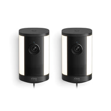 Pair of black Ring spotlight security cameras with integrated white side lights, front lenses, and cords, shown against a white background.