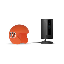 Black Ring indoor camera on a stand next to an orange mini football helmet with a bold “B” logo on white background.