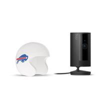 Black Ring indoor camera on stand next to a small white football helmet with a blue buffalo logo and red stripe on a white background.