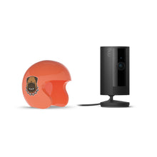 Orange open-face helmet with a bulldog sticker next to a black Ring indoor camera on a stand with power cable. White background.