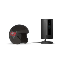 Black Ring indoor camera on a stand next to a small black football helmet with a red pirate-flag logo on white background.