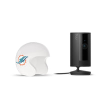Black Ring indoor camera on a stand beside a white mini football helmet with a teal dolphin logo, on a white background.