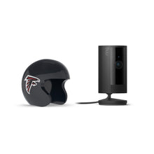 Black Ring indoor camera on a stand beside a dark football helmet with a red and white falcon logo on white background.