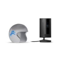 Black Ring indoor camera on a stand next to a gray helmet with a blue lion logo, set against a white background.
