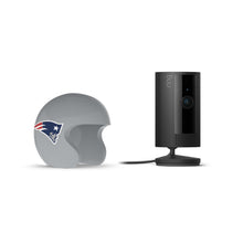Black Ring indoor camera on a stand beside a gray football helmet with a Patriots logo, set against a white background.