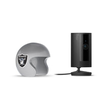 Black Ring indoor camera on a stand next to a silver football helmet with the Raiders logo, on a white background.