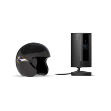 Black Ring indoor camera on a stand beside a glossy black football helmet with a purple-and-gold raven logo, set against a white background.