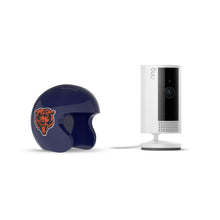 White Ring indoor camera on a stand beside a navy mini football helmet with an orange bear logo on a plain white background.