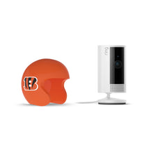 White Ring indoor camera on a stand next to an orange mini football helmet with a black “B” logo, on a white background.