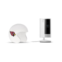 White Ring indoor camera on a stand beside a white mini football helmet with a red cardinal logo, on a plain white background.