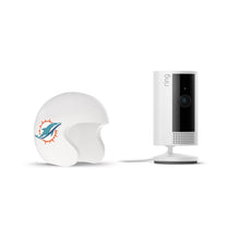 White Ring indoor camera on a stand next to a white mini football helmet with a teal dolphin logo on the side, set against a plain background.