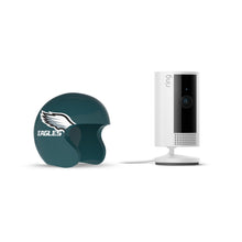 White Ring indoor camera on a stand next to a dark green mini helmet with the Eagles wing logo on a white background.