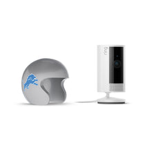 White Ring indoor camera on a stand beside a silver mini football helmet with a blue lion logo on a white background.