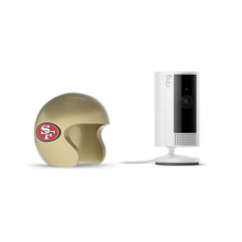 Ring indoor camera on a stand beside a gold football helmet with the San Francisco 49ers logo, set against a white background.