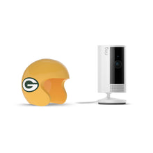 White Ring indoor camera on a stand beside a yellow Green Bay Packers mini helmet on a white background.