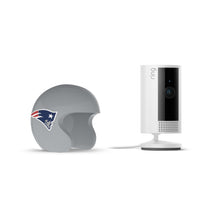White Ring indoor camera on a stand beside a gray football helmet with the New England Patriots logo on a white background.