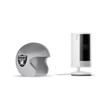White Ring indoor camera on a stand beside a glossy gray Raiders-themed mini helmet on a white background.