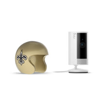 Gold football helmet with a black fleur-de-lis next to a white Ring indoor camera on a stand with power cable, set against a white background.
