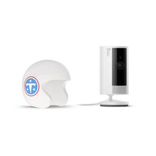 White Ring indoor camera on a stand next to a white helmet featuring a blue and red logo with stars on a white background.