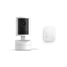 White Ring indoor camera on a stand with black front panel and lens, shown beside a small white Ring sensor on a plain background.