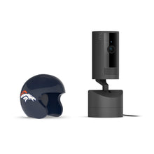 Black Ring indoor camera on a stand next to a dark blue football helmet with a white horse logo, set against a white background.