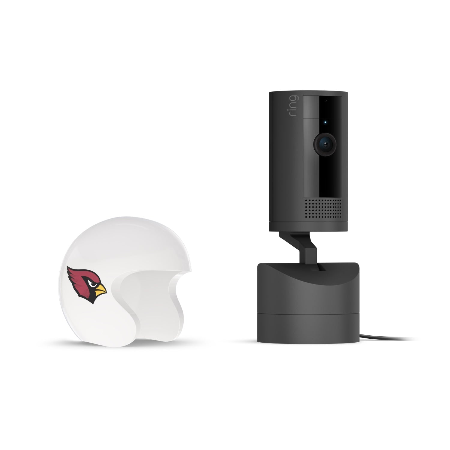 Black Ring indoor camera on a stand next to a small white helmet with a red cardinal logo, on a plain white background.
