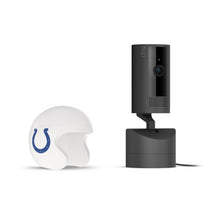 Black Ring indoor camera on a stand next to a white helmet with a Colts logo on a plain white background.