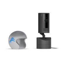 Black Ring indoor camera on a base next to a gray mini football helmet with a blue lion logo, set against a white background.