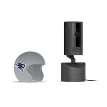 Black Ring indoor camera on a base next to a gray mini football helmet with the New England Patriots logo on a white background.