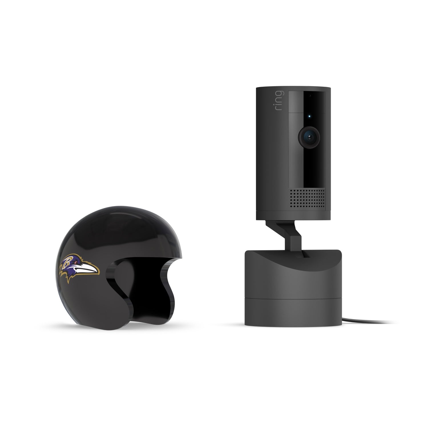Black Ring indoor camera on a charging base beside a glossy black mini helmet with a purple raven logo on a white background.