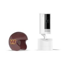 White Ring indoor camera on a stand next to a maroon mini football helmet with a yellow “W” logo on a white background.