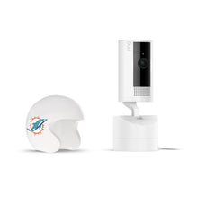 White Ring indoor camera on stand next to a white mini football helmet with a teal dolphin logo on a plain white background.