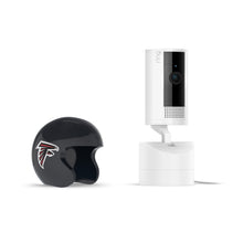 White Ring indoor camera on a stand next to a small black Atlanta Falcons helmet on a white background.