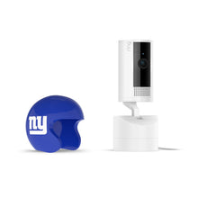 White Ring indoor camera on a stand next to a blue mini football helmet with white “ny” logo on a plain white background.