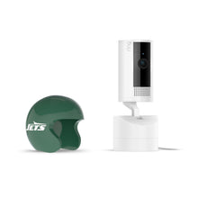 White Ring indoor camera on a stand beside a green Jets mini helmet cover on a plain white background.
