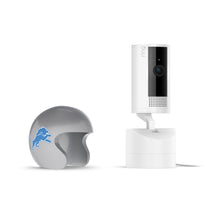 White Ring indoor camera on stand with power cable, next to a gray mini football helmet featuring a blue lion logo on a white background.