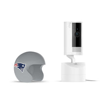 White Ring indoor camera on a charging dock beside a gray mini football helmet with the New England Patriots logo on it.