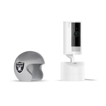 White Ring Pan-tilt indoor camera on stand beside a gray Raiders mini helmet with team logo, set against a clean white background.