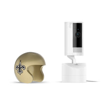 White Ring indoor camera on a stand next to a small gold helmet with a black fleur-de-lis logo on a white background.