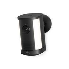 Wall-mounted black Ring camera with dual white light panels, wide lens, and speaker grille on a round base against a white background.