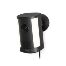 Black Ring wall-mounted security camera with wide LED light panels, lens centered, small indicator light, and power cord attached.